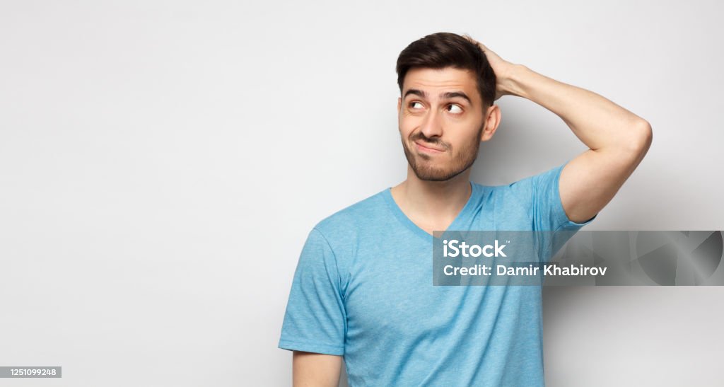 Young doubtful man thinking, scratching head and trying to find solution, isolated on gray background Uncertainty Stock Photo