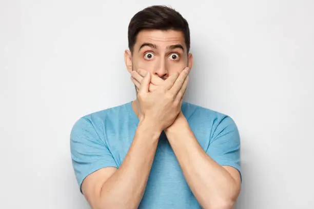 Shocked man dressed in blue t-shirt, covering mouth with both hands, eyes round with shock and fear, isolated on gray background