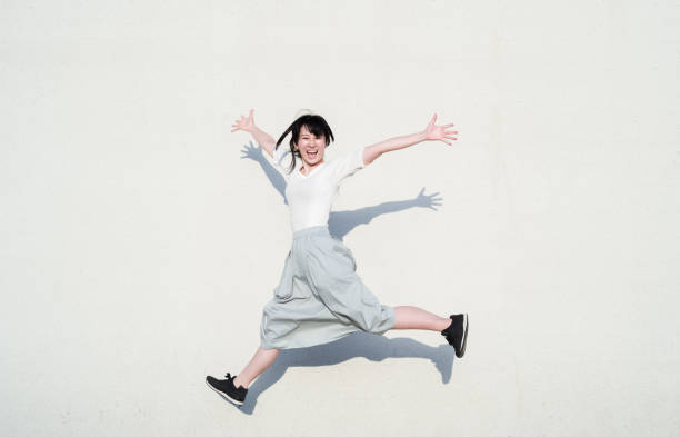 foto de mulher asiática pulando com sorriso em frente a parede branca - beautiful individuality joy enjoyment - fotografias e filmes do acervo