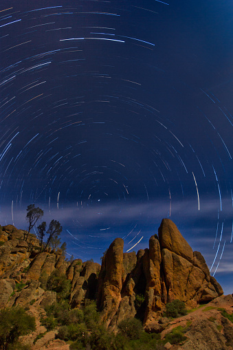 Star trails in a circular pattern in a dark night sky in California. Brown rocks and trees in the foreground.