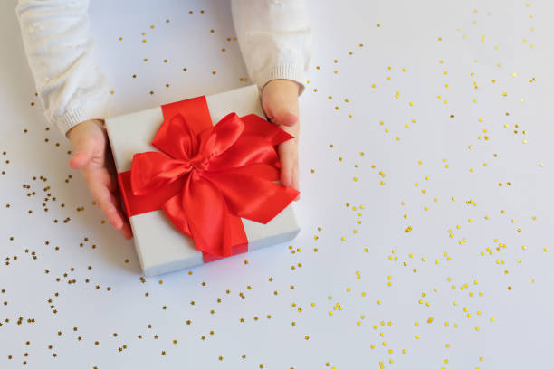 istock Baby hands holding present with red bow on light background with gold sparkles. 1251107227