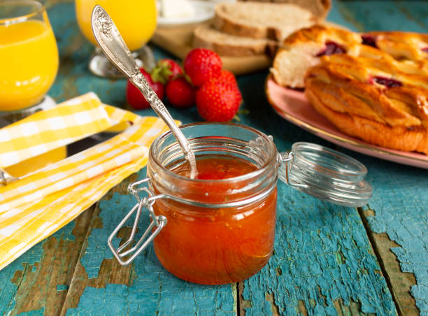 Apricot jam in a jar with a silver spoon on the table at breakfast. Two glasses with orange juice, cherry pie, ripe strawberries, butter knife, napkin on the background."n