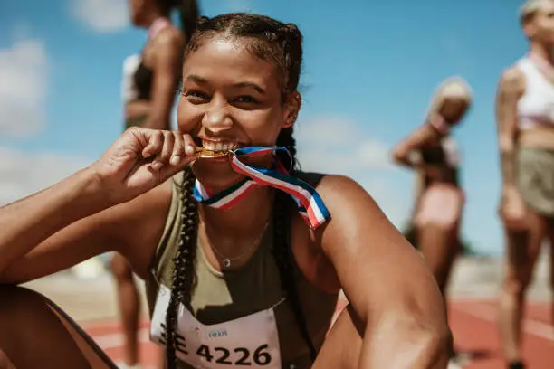Excited female athlete biting her medal while sitting on race track with other athletes in background. Sports woman enjoying winning a medal in sprinting event at stadium.