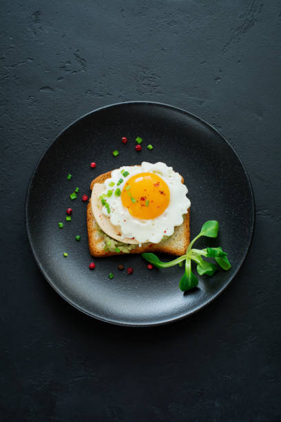 istock Toast with guacamole sauce from avocado, cheese, fried egg and fresh on black ceramic plate on dark concrete background. Selective focus. Top view. 1251142576