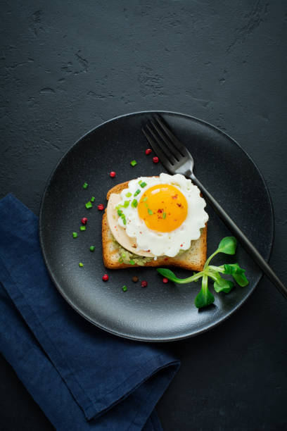 istock Toast with guacamole sauce from avocado, cheese, fried egg and fresh on black ceramic plate on dark concrete background. Selective focus. Top view. 1251143252