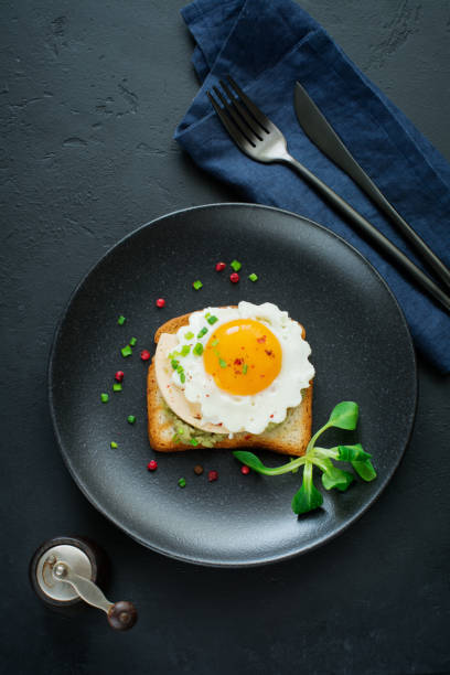 istock Toast with guacamole sauce from avocado, cheese, fried egg and fresh on black ceramic plate on dark concrete background. Selective focus. Top view. 1251143479