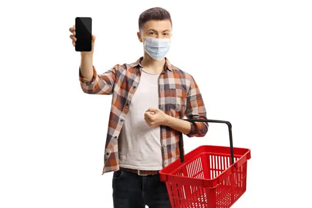Young man with a protective face mask and an empty shopping basket showing a mobile phone to the camera isolated on white background