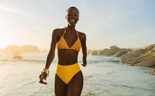 Attractive african woman in yellow bikini walking along the beach. Smiling female in swimwear coming out of the sea water at sunset.