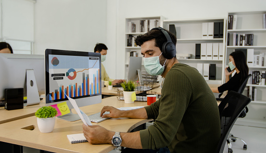 social distancing concept, business man wearing mask, working and looking at finance graph paper and monitor in office, sit far away from other people