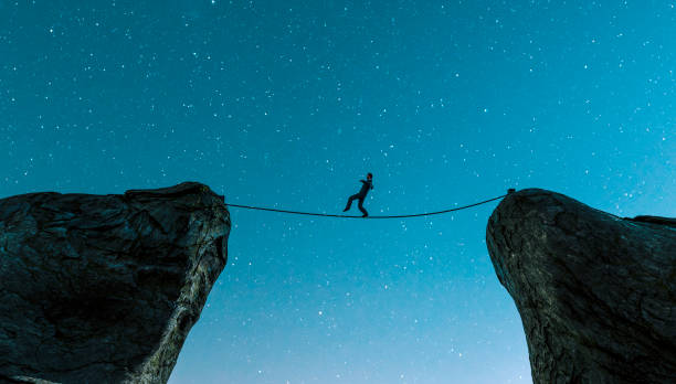 Walking on rope between two cliffs and keeping the balance Man walks carefully on a rope that has been tied between two cliffs. He risks falling to the ground. Concept of success and avoiding problems following a career path. 
Note: The man is a 3D-render. Model release attached. tightrope stock pictures, royalty-free photos & images