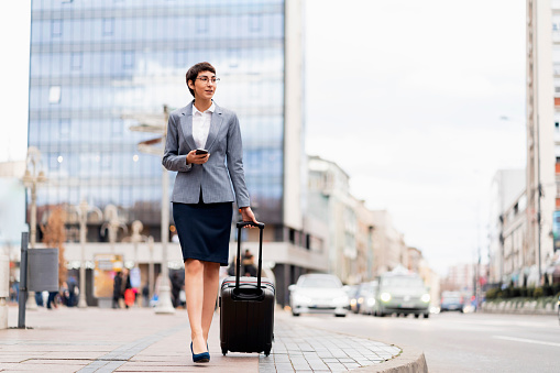 Young Caucasian businesswoman arrived at her destination. Female entrepreneur on a business trip. She is holding a phone in her hand and dragging a suitcase.