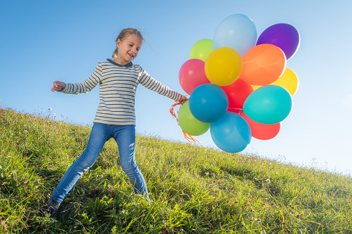 Cheerful little girl standing on a meadow and playing with a cluster of colorful balloons.