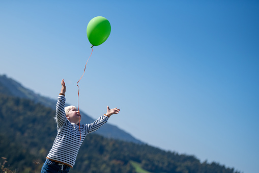Joyful blond caucasian boy releasing a green balloon into the sky. Hills and sky in the background.