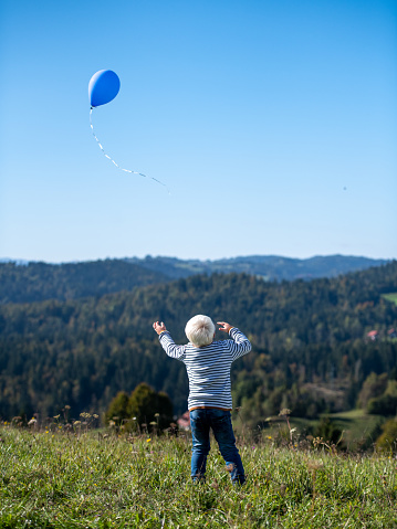 Blond boy standing on a meadow on a hill releasing a balloon into the sky.