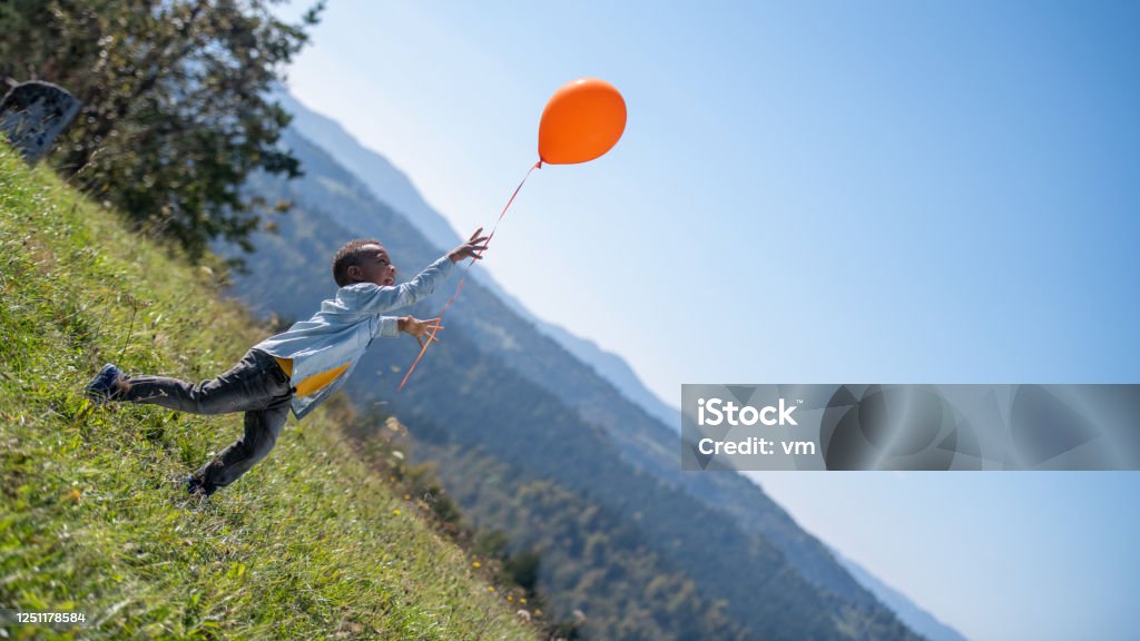 Boy on a meadow releasing an orange balloon Little boy of African descent standing on a meadow in nature and releasing an orange balloon into the sky. Hills and sky in the background. Balloon Stock Photo