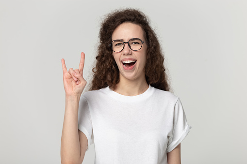 Happy teenage girl punk in white t-shirt and glasses isolated on grey studio background look at camera show rock-n-roll hand gesture, smiling crazy millennial female pose with heavy metal horns sign