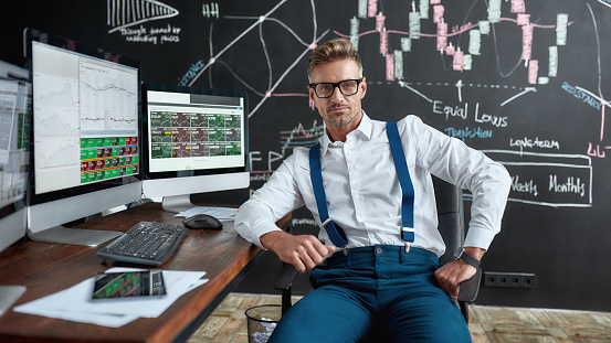 Middle-aged caucasian trader in glasses looking at camera while sitting by desk in front of computer monitor. Blackboard full of charts and data analyses in background. Stock trading, people concept.