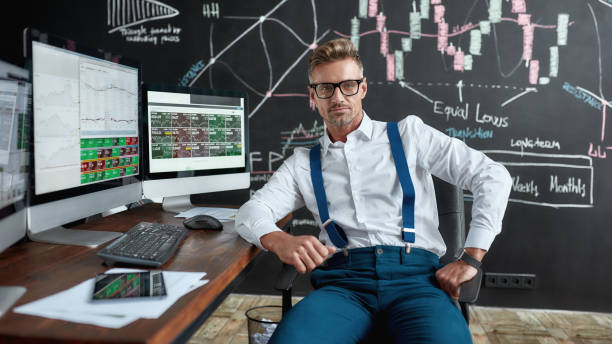 Letting money grow. Middle-aged caucasian trader in glasses looking at camera while sitting by desk in front of computer monitor. Blackboard full of charts and data analyses in background. Middle-aged caucasian trader in glasses looking at camera while sitting by desk in front of computer monitor. Blackboard full of charts and data analyses in background. Stock trading, people concept. stock certificate growth price market stock pictures, royalty-free photos & images