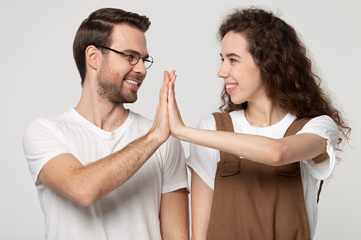 Smiling young man and woman isolated on grey studio background look in eyes give high five celebrate shared win, happy millennial couple join hands show love support and care, promise unity