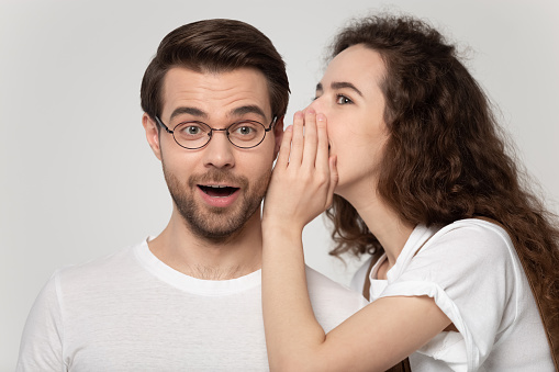 Millennial teenage girl whisper in surprised boyfriend ear tell secret about good sale offer or promotion, young couple isolated on grey studio background share private hidden information or data