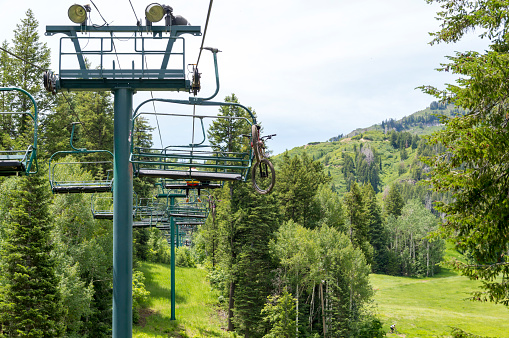This shows a ski lift at resort with a mountain bike hanging from one of the chairs.  The bike's rider is in the chair in front. This lift is popular with mountain bike riders in the summer.  Mountain bikes are hoisted up the lifts along with riders and a large number of downhill trails are available for the descent.