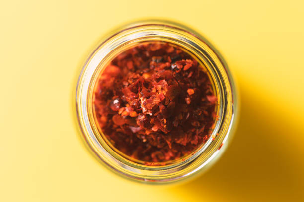 Chilli pepper flakes in glass jar on yellow background. Spices from directly above, selective focus.