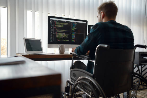 istock Multitasking. Back view of young caucasian male web developer in a wheelchair writing program code while sitting at workplace with three monitors in the modern office 1251204362
