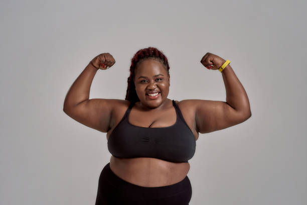 Enjoy your body. Plump, plus size african american woman in sportswear smiling at camera, showing strong arms in studio over grey background. Concept of sport, healthy lifestyle, body positive Plump, plus size african american woman in sportswear smiling at camera, showing strong arms in studio over grey background. Concept of sport, body positive, equality. Horizontal shot. Front view huge black woman pictures stock pictures, royalty-free photos & images