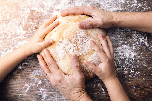 hands of mother and daughter touching a loaf of freshly baked rustic bread illuminated by the morning sun on a wooden table, concept of family and healthy food at home