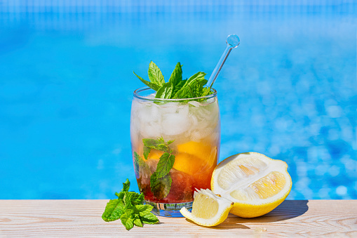 A fresh summer drink by the pool. The drink with mint leaves and lemon wedges is red at the bottom, and it has with crushed ice and fresh mint leaves at the top. Blue pool background, with room for text copy space.