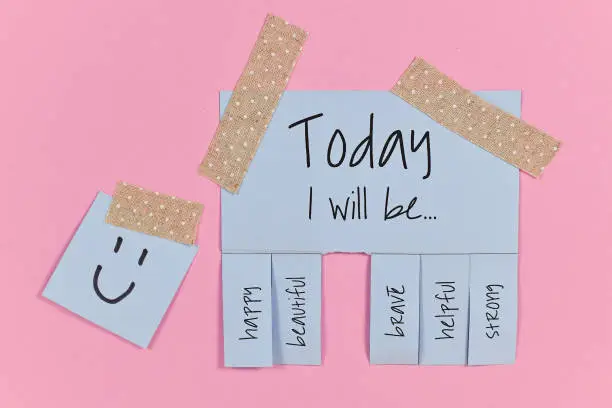 Motivational concept with light blue tear-off stub note with text 'Today I will be...' and words 'happy, beautiful, brave, helpful' and 'strong' and one stub missing on pink background
