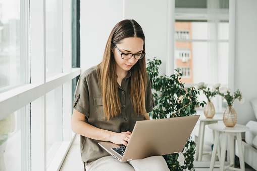 Beautiful young adult positive girl uses a laptop at home by the window or is she in open space.