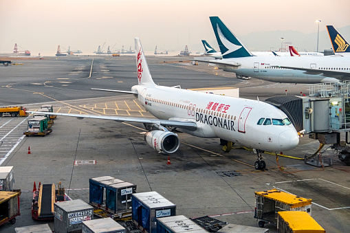 Hong Kong, China - Dec 11, 2019: Dragonair B-HSG (Airbus A320-232) being serviced and readied for its next flight at Hong Kong International Airport. In the foreground are a range of service equipment and in the background, are the tails of Cathay Pacific (including B-HNW, Boeing 777-31H) and Singapore Airlines planes plus a range of industrial barges moored in the foggy bay
