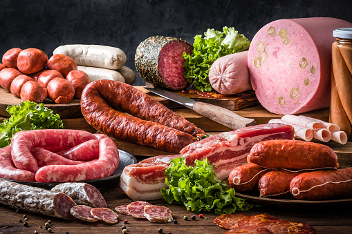 Front view of various kinds of raw sausages like mortadella, bacon, salami, ham and pickled sausages on a delicatessen concept background. Sausages are on a rustic wooden table. Studio shot taken with Canon EOS 6D Mark II and Canon EF 24-105 mm f/4L