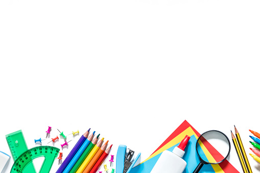 Top view of various kinds of multicolored school supplies like scissors, crayons, rulers, pencils, papers, markers, calculators, magnifying glass, clips, thumbtacks, sharpeners and brushes standing at the lower part of the image on a frame shape with copy space at the top of the image on a white background. Studio shot taken with Canon EOS 6D Mark II and Canon EF 24-105 mm f/4L