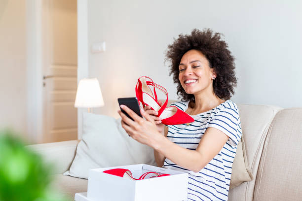Smiling woman unboxing a postal parcel and taking a selfie with her new purchases using a smartphone. Showing shoes to her friends via video call