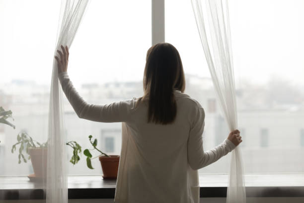 Photo of Rear view woman standing near window opening curtains looking outside
