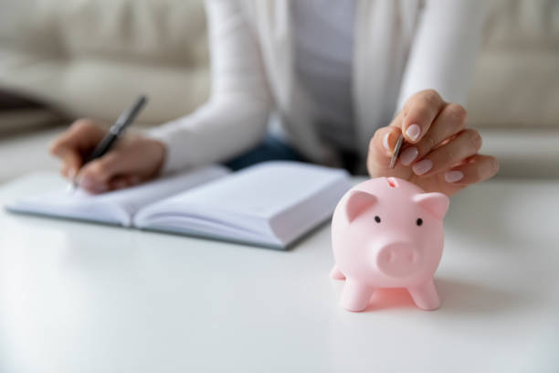 Thrifty woman writing daily expenses put coin into piggy bank Thrifty woman sit at table hold pen writing daily expenses in diary put coin in pink piggy bank close up. Saving money for future, caring for tomorrow, makes stash of cash, investment, economy concept inexpensive stock pictures, royalty-free photos & images