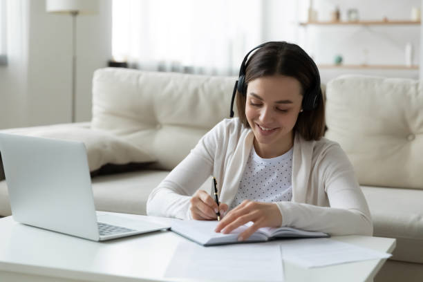 Smiling woman wear headset watch webinar writing notes Smiling woman wear headset watch webinar makes video conference on-line call writing notes. E-study via internet with teacher, student learn at home use laptop and headphones, customer service concept business teaching student writing stock pictures, royalty-free photos & images