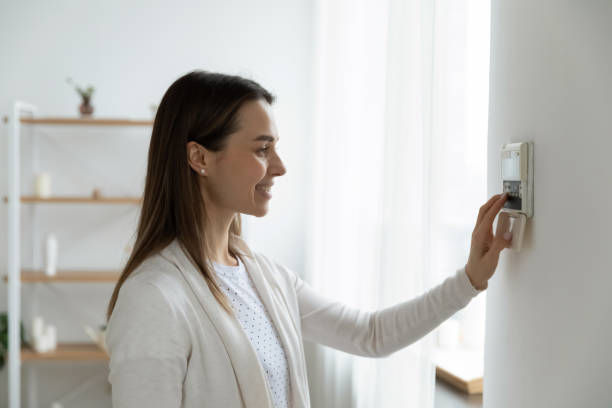 Smiling woman adjusting degrees set comfortable temperature using thermostat Smiling woman standing near wall-mounted device adjusting degrees in living room set comfortable temperature using thermostat home heating system. Owner of modern smart house, energy saving concept home heating stock pictures, royalty-free photos & images