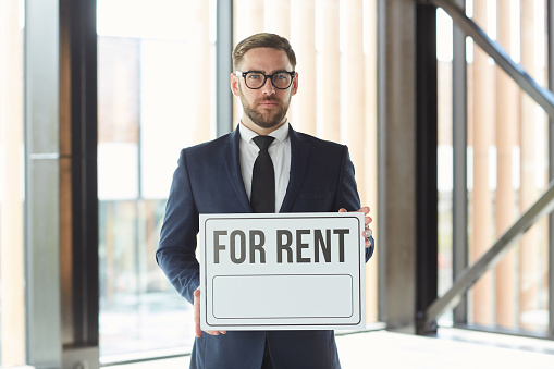 Portrait of mature businessman in suit holding placard For Rent and looking at camera while standing at modern office