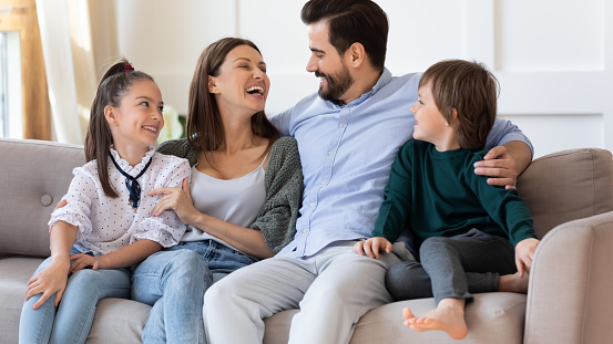 Happy mother and father hugging with children, embracing, laughing, sitting on couch, family having fun at home, parents with adorable son and daughter spending leisure time on weekend together