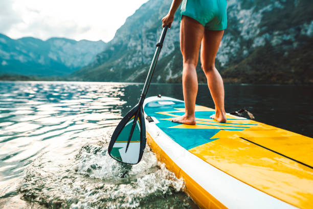 Woman in Early 30s Paddleboarding on Lake Bohinj in Slovenia Low angle view of fit mid adult woman moving past camera on paddleboard as she explores lake in Triglav National Park with Julian Alps in background. using a paddle stock pictures, royalty-free photos & images