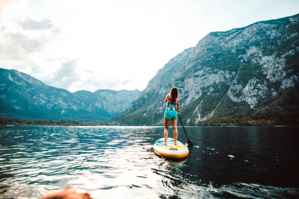 Full length front view of woman in early 30s smiling and looking away from camera as she oars paddleboard on Lake Bohinj in Triglav National Park, Slovenia.