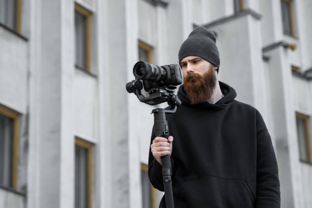 istock Bearded Professional videographer in black hoodie holding professional camera on 3-axis gimbal stabilizer. Filmmaker making a great video with a professional cinema camera. Cinematographer. 1251297532