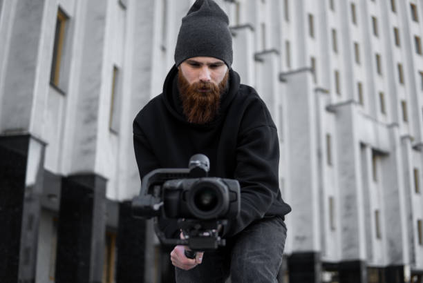 istock Bearded Professional videographer in black hoodie holding professional camera on 3-axis gimbal stabilizer. Filmmaker making a great video with a professional cinema camera. Cinematographer. 1251297652