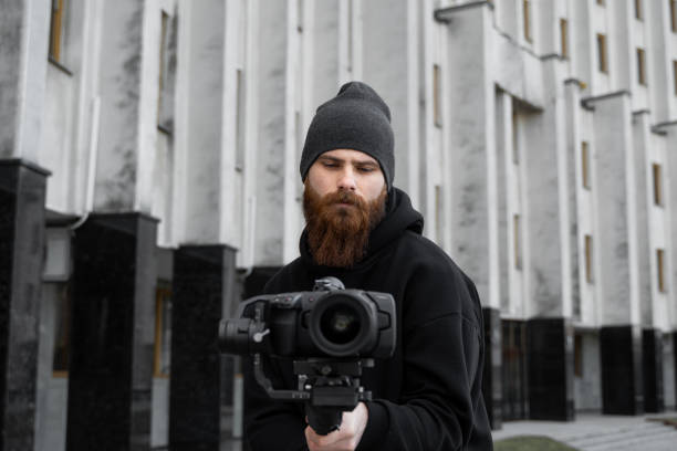 istock Bearded Professional videographer in black hoodie holding professional camera on 3-axis gimbal stabilizer. Filmmaker making a great video with a professional cinema camera. Cinematographer. 1251299100