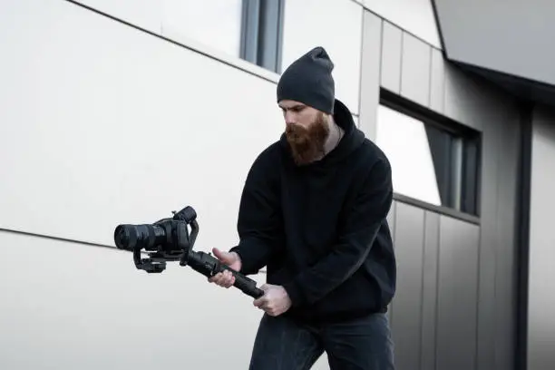 Bearded Professional videographer in black hoodie holding professional camera on 3-axis gimbal stabilizer. Filmmaker making a great video with a professional cinema camera. Cinematographer