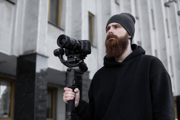 istock Bearded Professional videographer in black hoodie holding professional camera on 3-axis gimbal stabilizer. Filmmaker making a great video with a professional cinema camera. Cinematographer. 1251299251