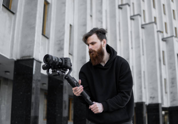 istock Bearded Professional videographer in black hoodie holding professional camera on 3-axis gimbal stabilizer. Filmmaker making a great video with a professional cinema camera. Cinematographer. 1251299286
